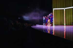 Fünf bunt verkleidete Personen stehen auf einer lila beleuchteten Bühne nebeneinander.