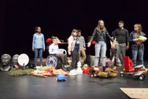 Eight young people are on a stage. One of them is sitting in a wheelchair. In front of them are many different objects such as a helmet, cuddly toys or a model aeroplane.