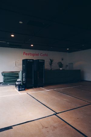 View of vending machines and a darkened counter of the Festspiel Café. The open floor in the foreground has been divided into black and white rectangles with tape.