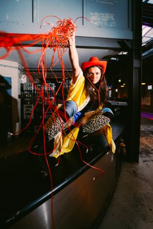 A female read person with a red cowboy hat and colourful as well as patterned clothes squats on a bar. She raises her hand vertically and swings red thin cables in the air.