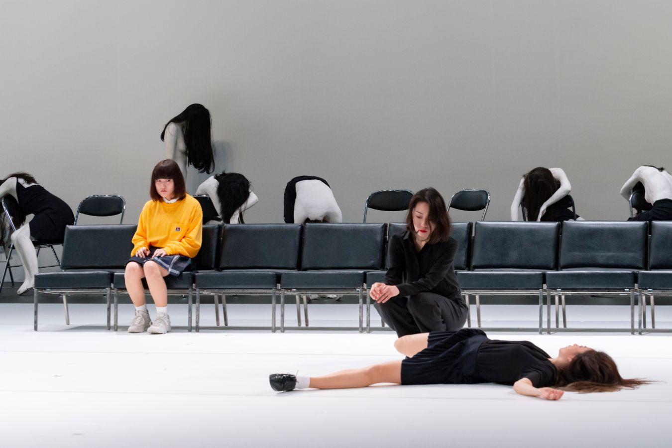 Eine Person in gelbem Pullover sitzt auf einer Stuhlreihe und schaut auf eine andere Person, die auf dem Boden liegt.