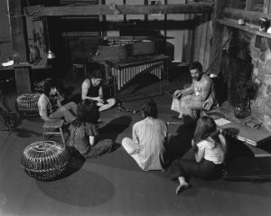 Six people are sitting on the floor of a room. In front of them and around them are instruments such as a saxophone, a vibraphone and a trumpet.