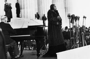 Marian Anderson steht in einem Pelzmantel vor einer Gruppe Menschen. Vor ihr befinden sich Mikrofone, direkt hinter ihr sitzt ein Mann an einem Flügel. Im Hintergrund sind monumentale Säulen erkennbar.