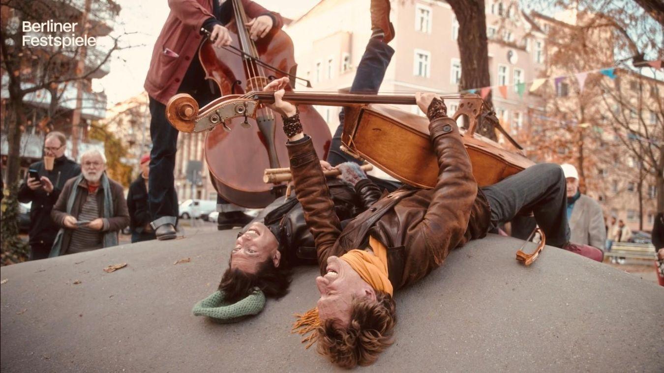 Zwei Männer liegen auf etwas gewölbtem. Einer hat ein Cello in der Hand. Eine weitere Person mit Cello steht neben ihnen.