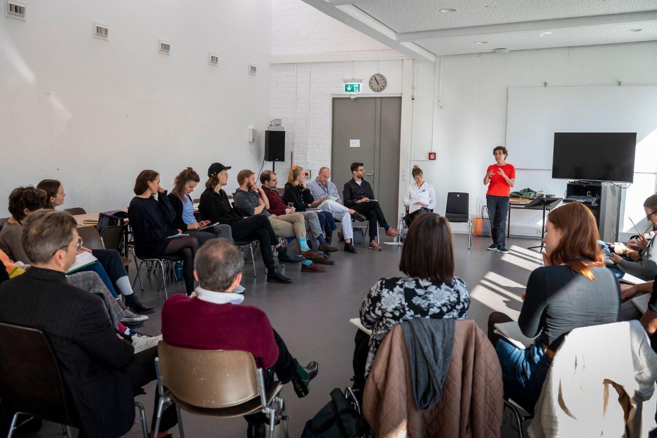 A woman in a red T-shirt speaks in front of a group of attentive listeners.