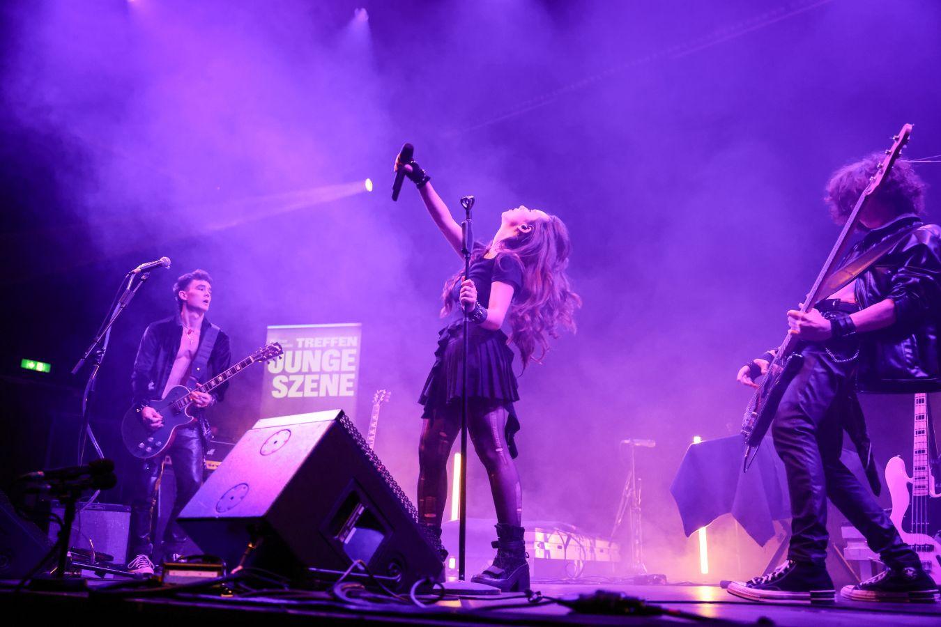 Three musicians are standing on a stage lit with purple lighting.