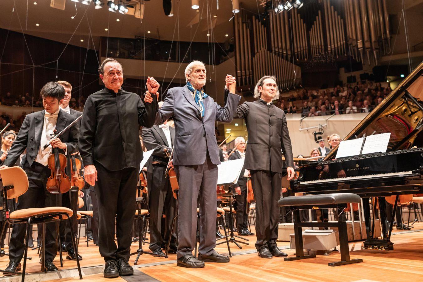 Drei Männer im Anzug stehen vor dem Orchester auf der Bühne und halten sich an den Händen.