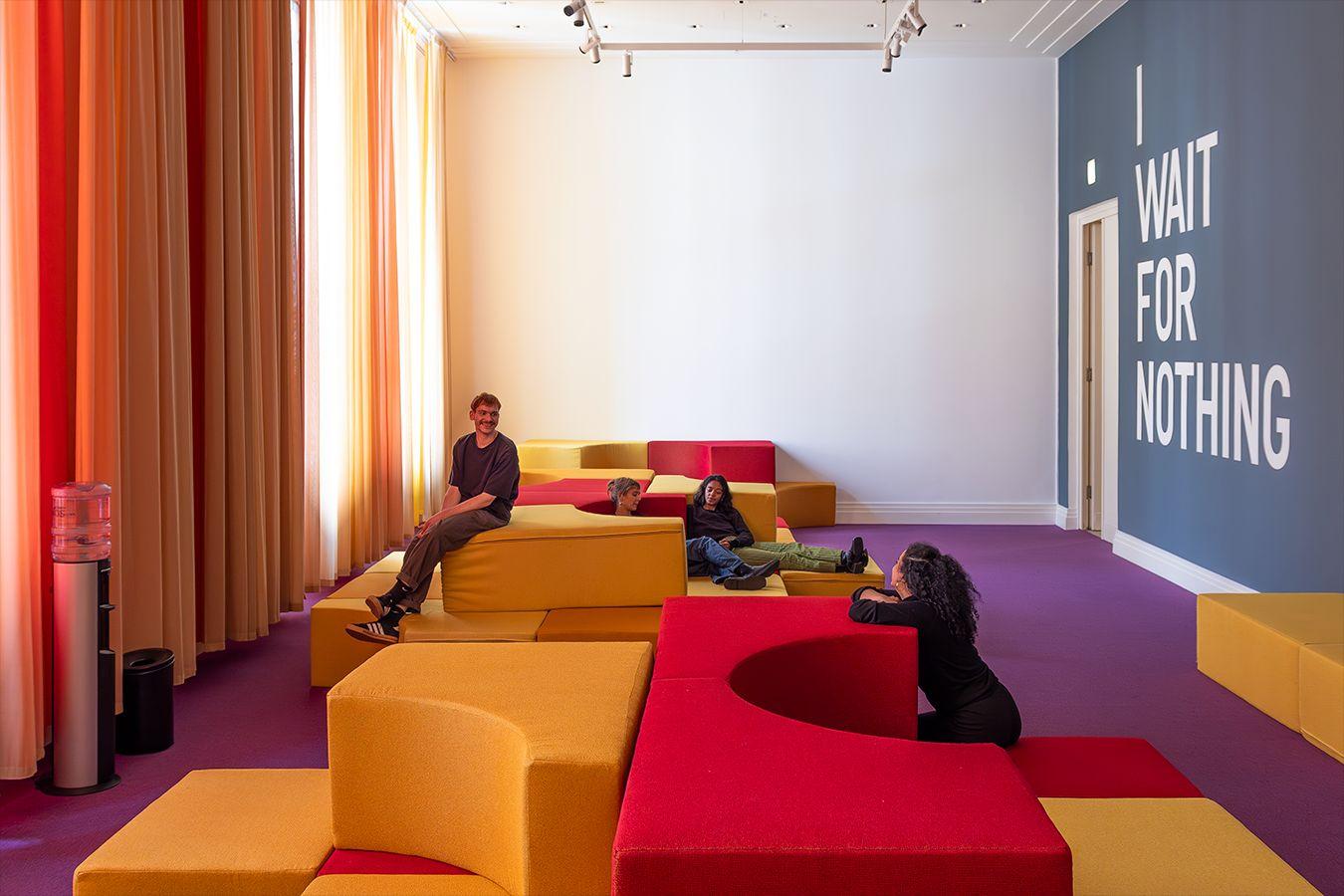 A shot of a room with a purple carpet, featuring various upholstered elements in yellow and red, with four people sitting on them. Along the length of the left wall hang yellow and red curtains, while the right wall is petrol-colored with a large white inscription reading 'I WAIT FOR NOTHING'.