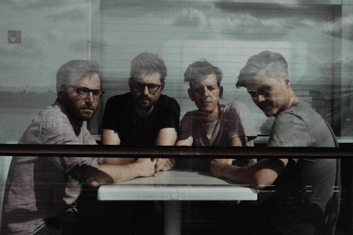 The four musicians of Nikel are sitting at a table. In front of them is a window that distorts the view.