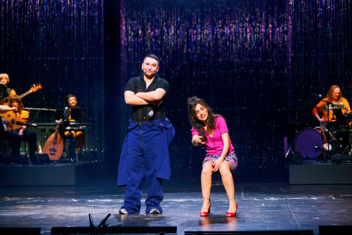 Ein Mann steht mit verschränkten Armen neben einer sitzenden Frau im pinken Kostüm, dahinter Menschen mit Instrumenten.