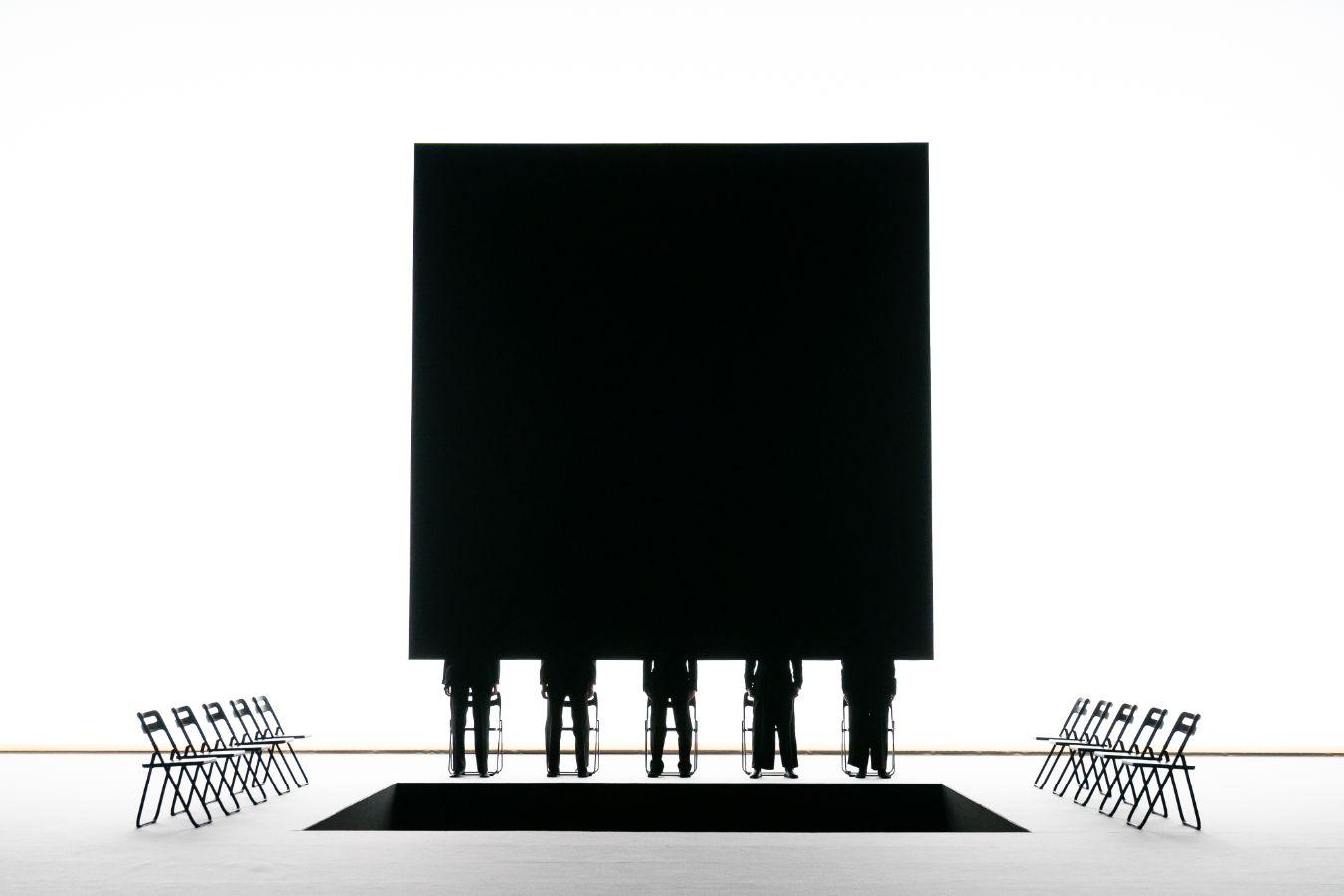 A white stage with a square opening, a large black screen behind it, and five black silhouettes in front.