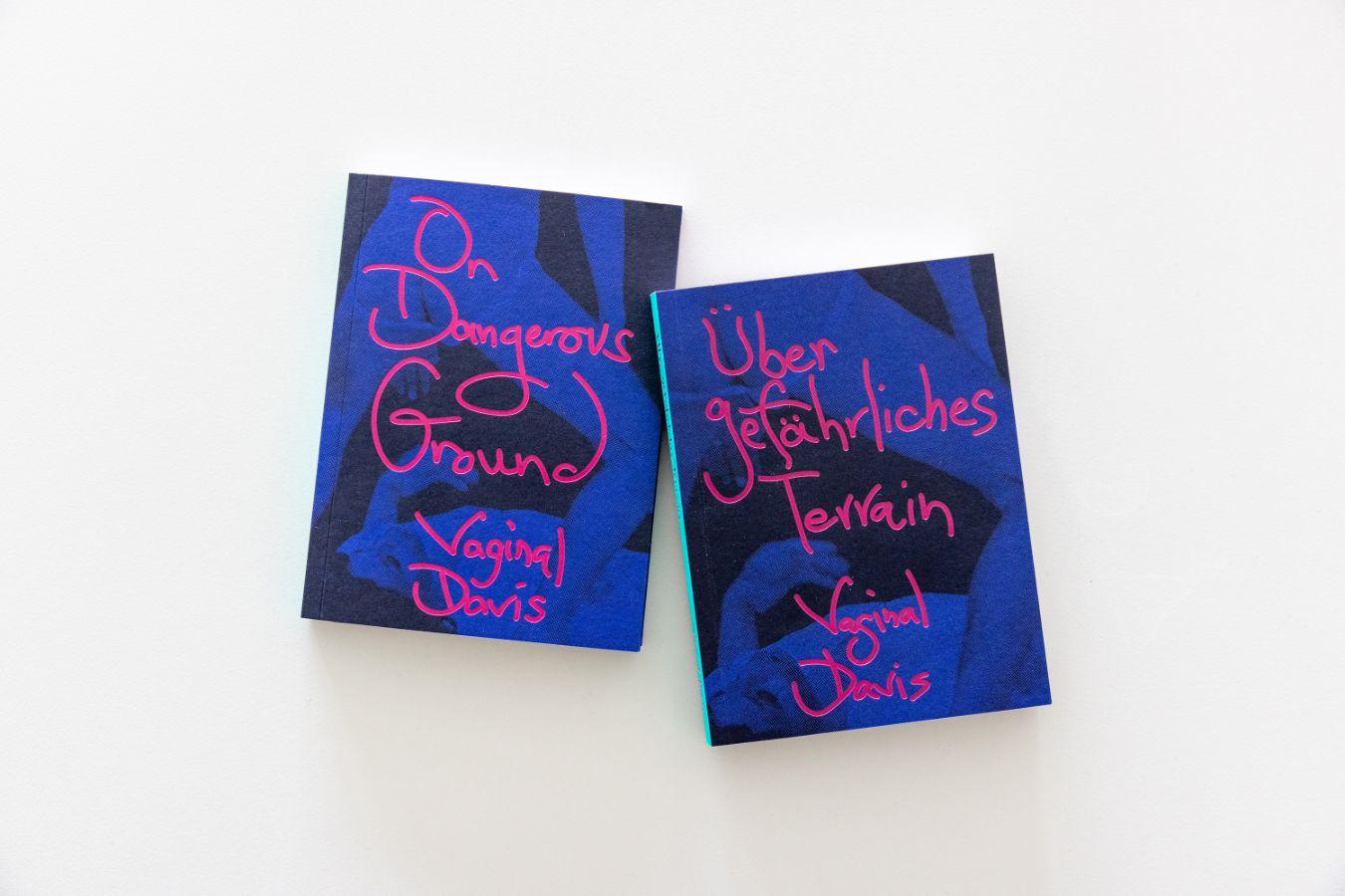 Zwei lilafarbene Bücher liegen auf hellen Hintergrund, auf ihnen steht in magenterfarbener Schrift gedruck "Über gefährliches Terrain. Vaginal Davis und "On Dangerous Ground. Vaginal Davis".
