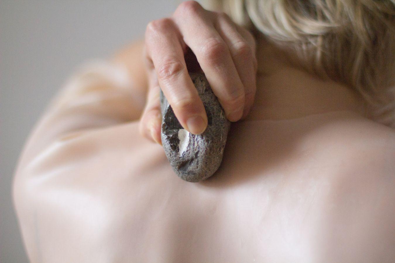 A person holds a stone in the hand and strokes her naked back with it. The cut-out shows the shoulder and lower head area.