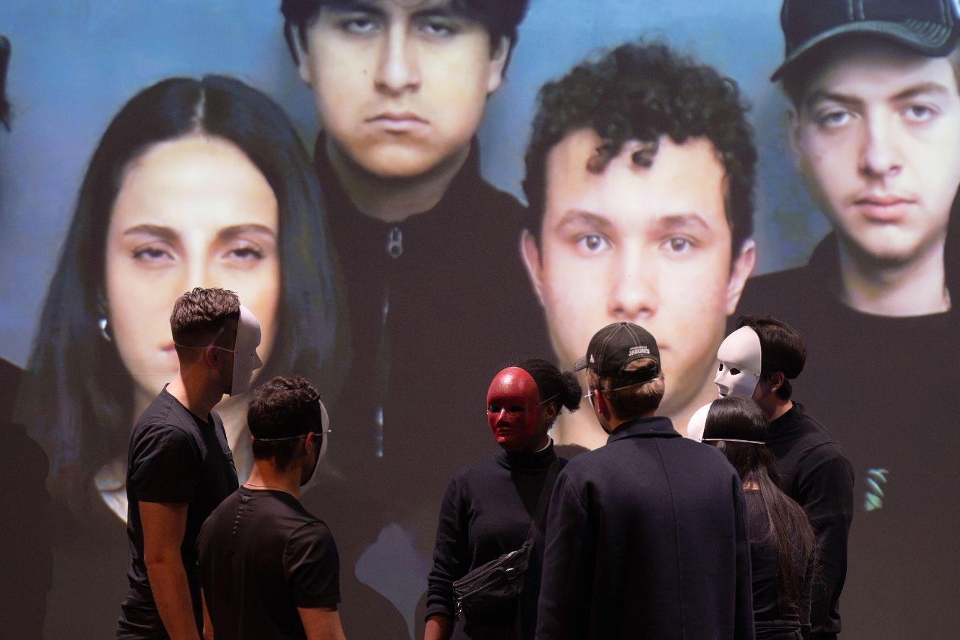 Pupils perform theatre, wearing masks, standing in a circle. In the background is a screen with their uncovered faces.