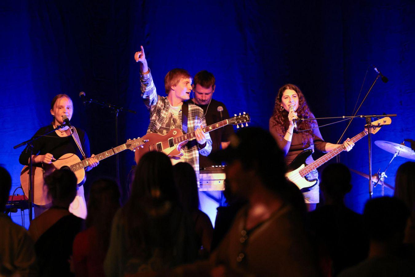 Four young musicians perform on a blue-lit stage with guitars and microphones.