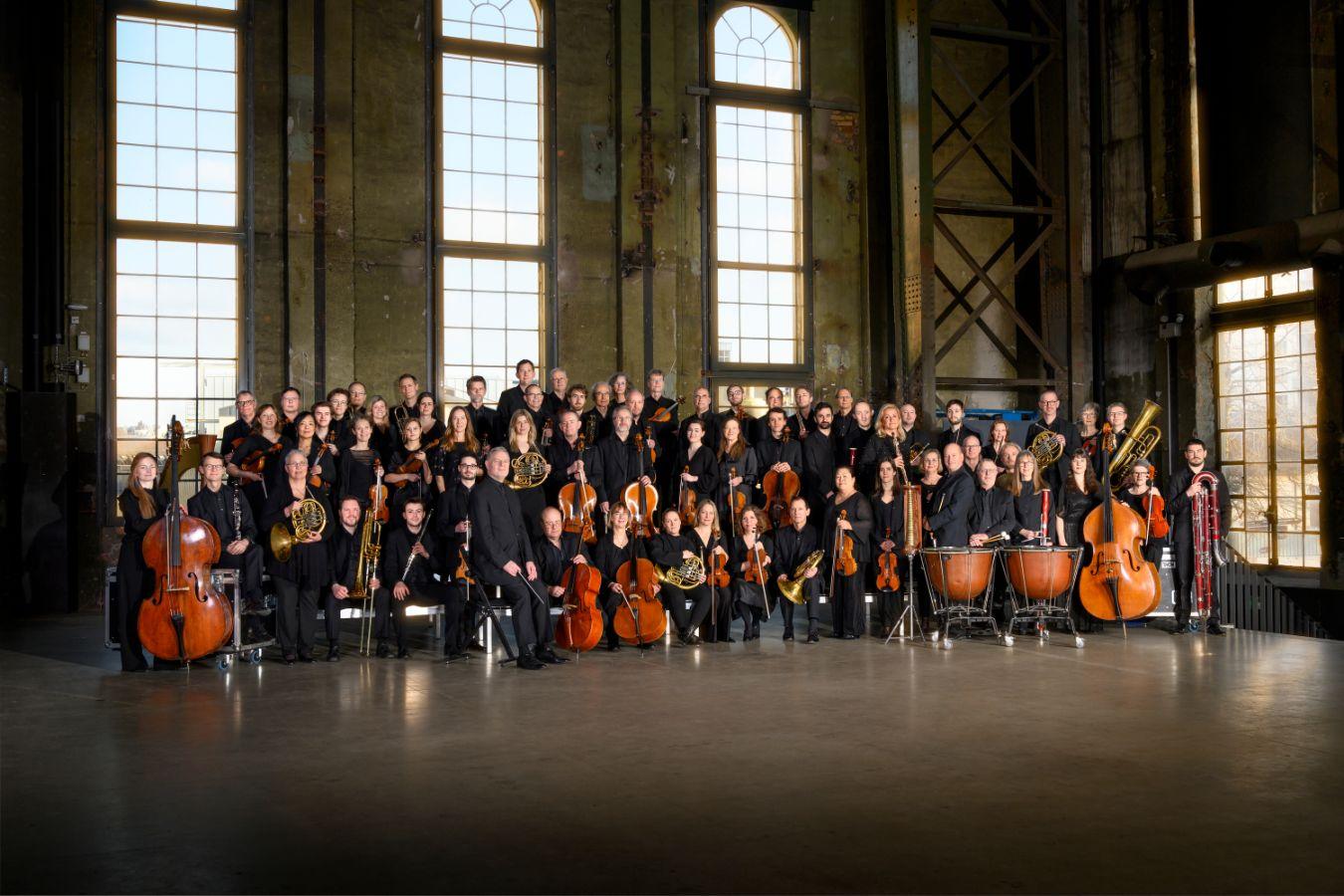 The orchestra stands in five long rows in front of a wall of a hall with large windows.