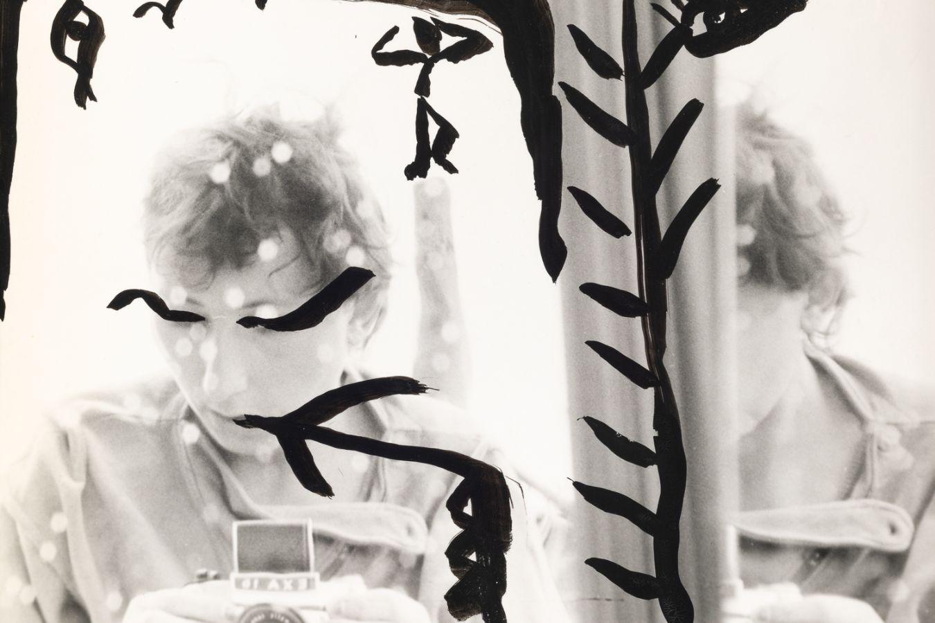 Ein mit schwarzen Mustern bemaltes Selbstporträt von Gabriele Stötzer, die sich mit einer Analogkamera im Spiegel fotografiert.