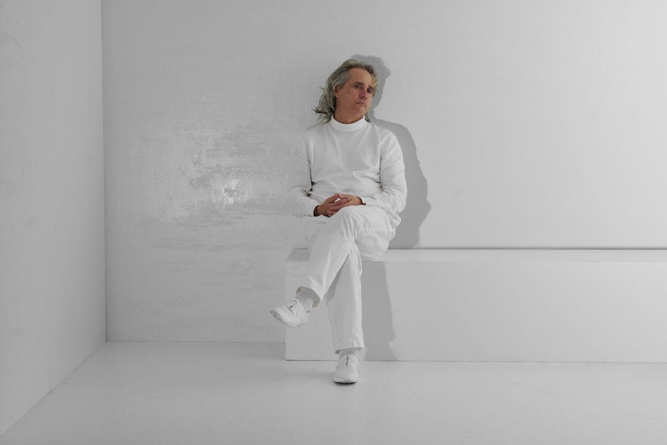 A man dressed in white sits with his legs crossed on a white bench inside a white cube