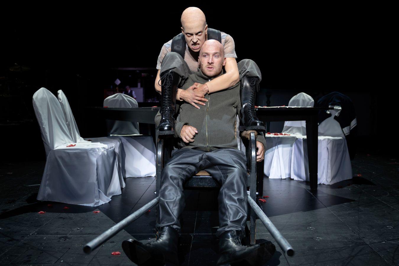 A bald person with an angry expression restrains a frightened‑looking man from behind