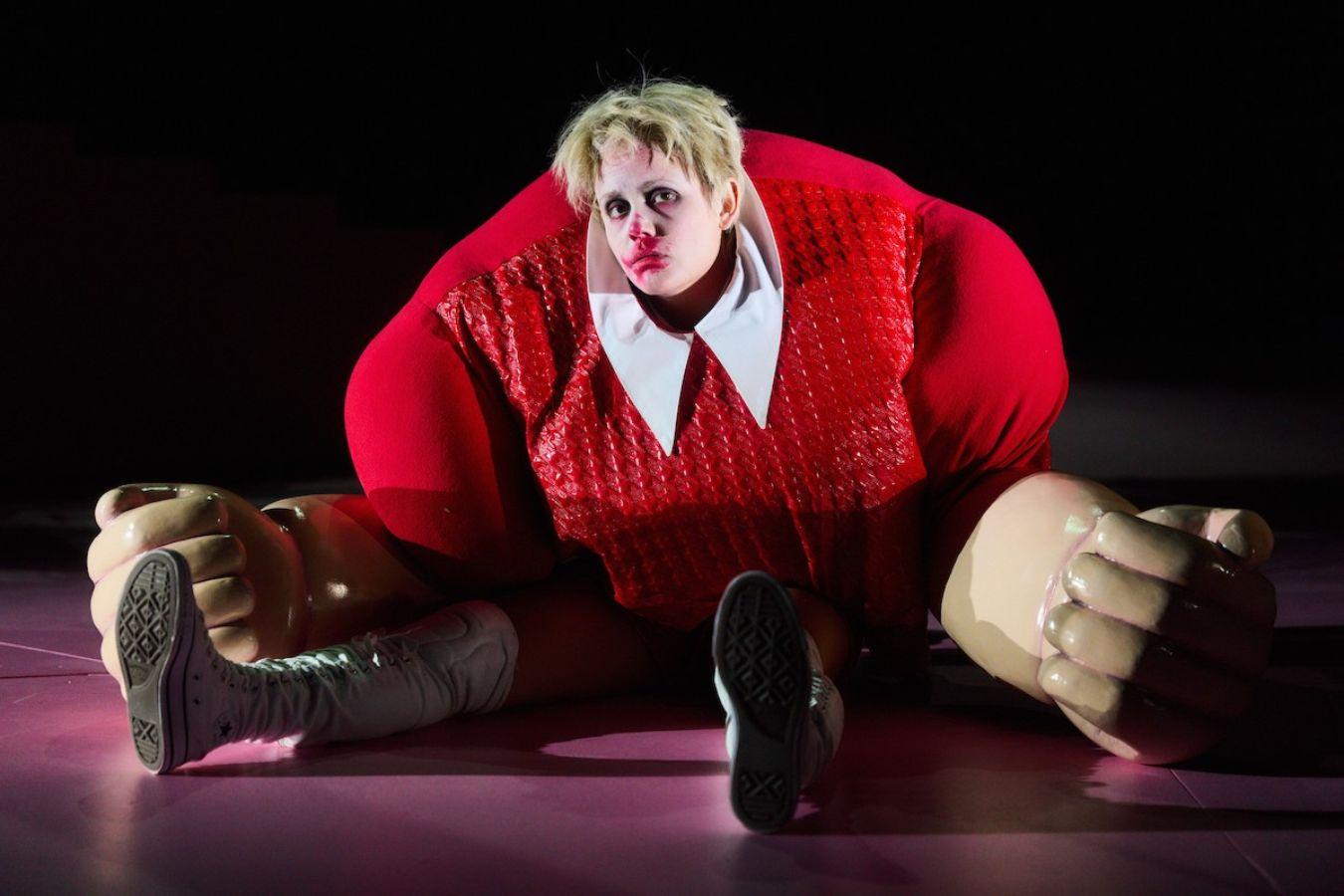 A woman in an oversized red costume with artificial fists sits on the floor with a blood‑smeared nose