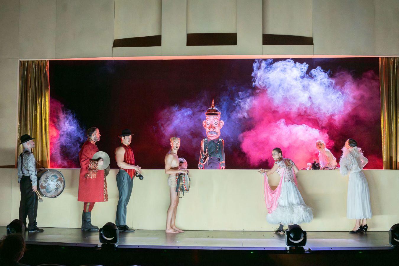 Men and women with instruments stand in front of a kind of puppet‑theatre booth featuring a uniformed figure with a papier‑mâché head