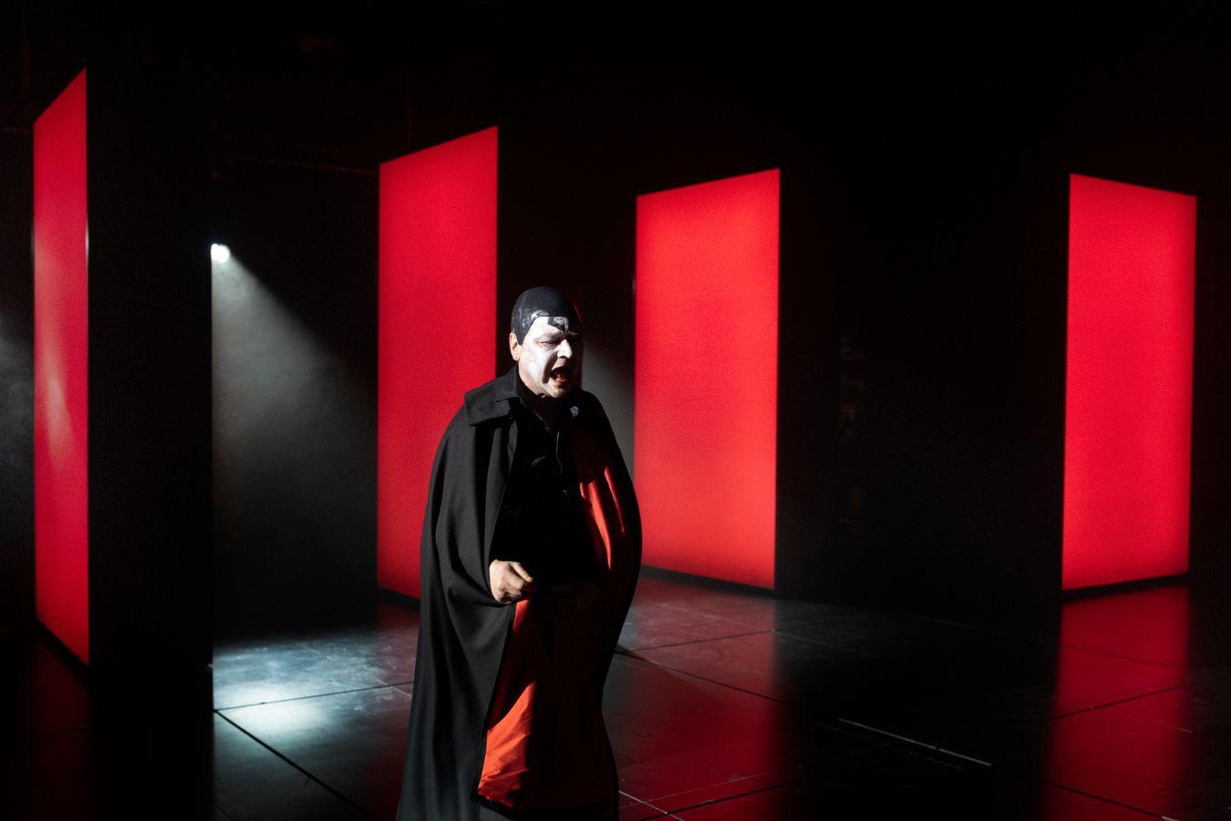 A man with white face paint and a red‑and‑black cloak stands in a red‑and‑black room