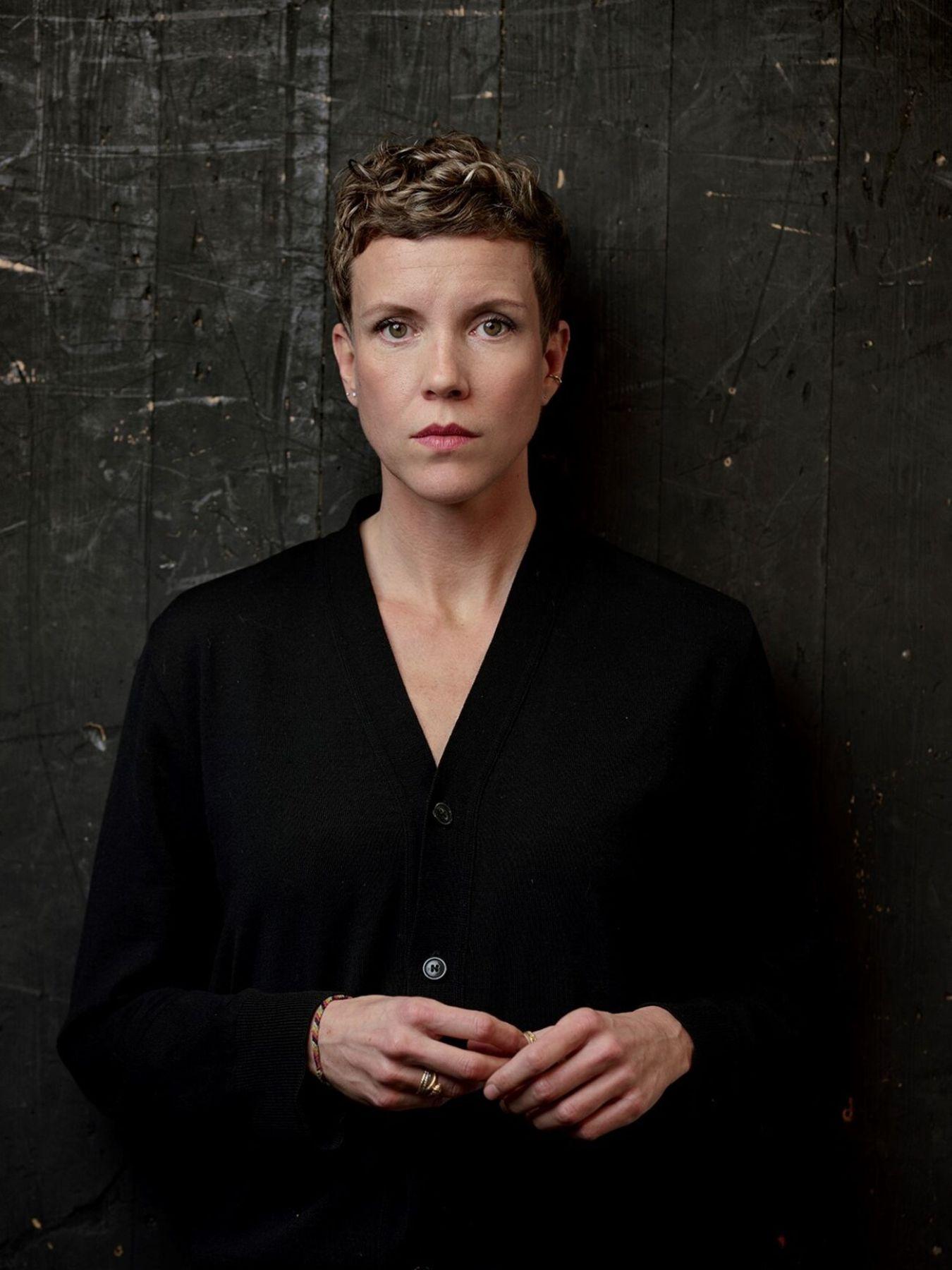 Person with short curly hair wearing a black buttoned shirt stands against a dark, textured wooden background, looking directly at the camera with a neutral expression.