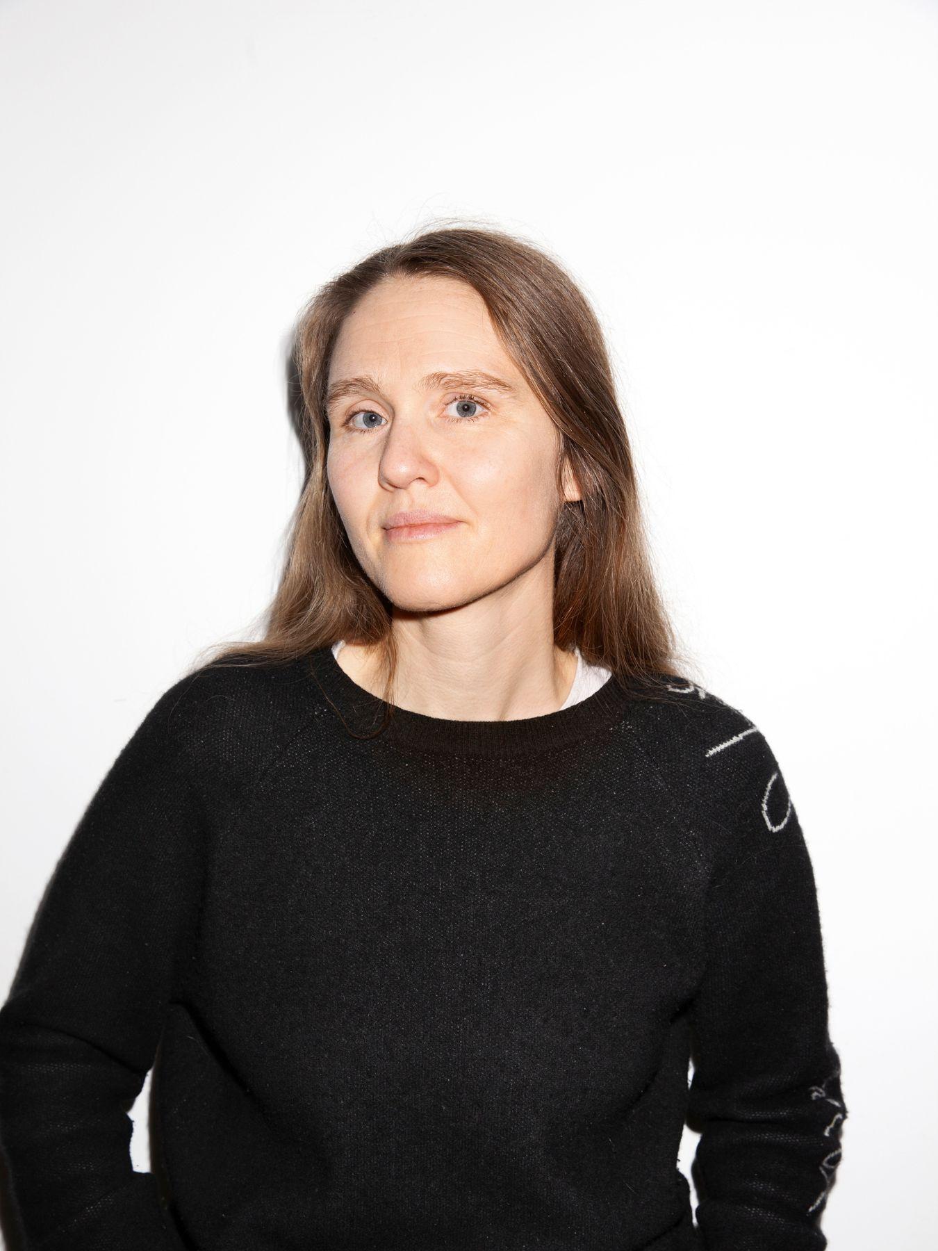 Person in dark sweater stands against a white wall with hands in pockets, upper body frontal.