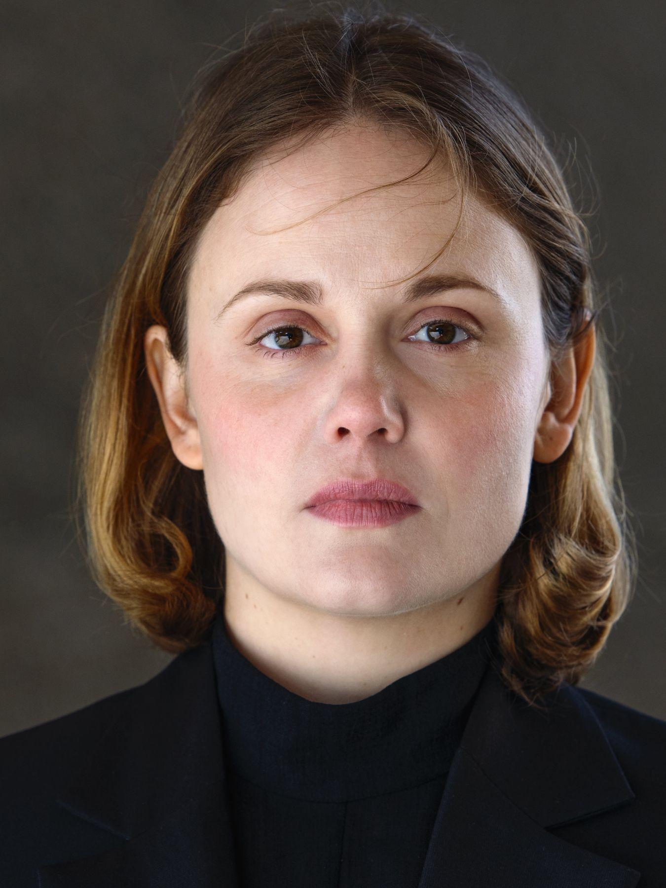 Studio portrait of a person against a dark background, wearing a black jacket with high collar, front view.