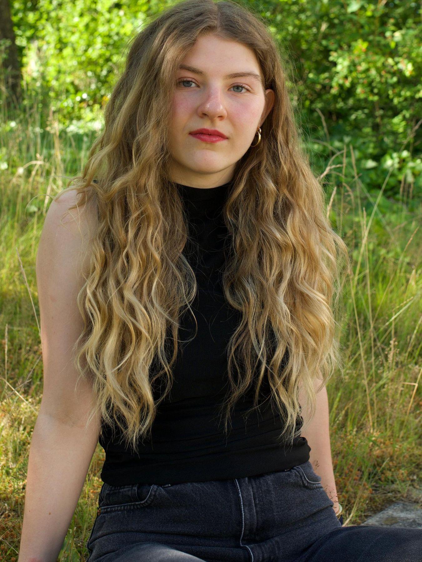 Person sitting on grass in a green outdoor area, wearing a dark top and jeans, surrounded by trees.