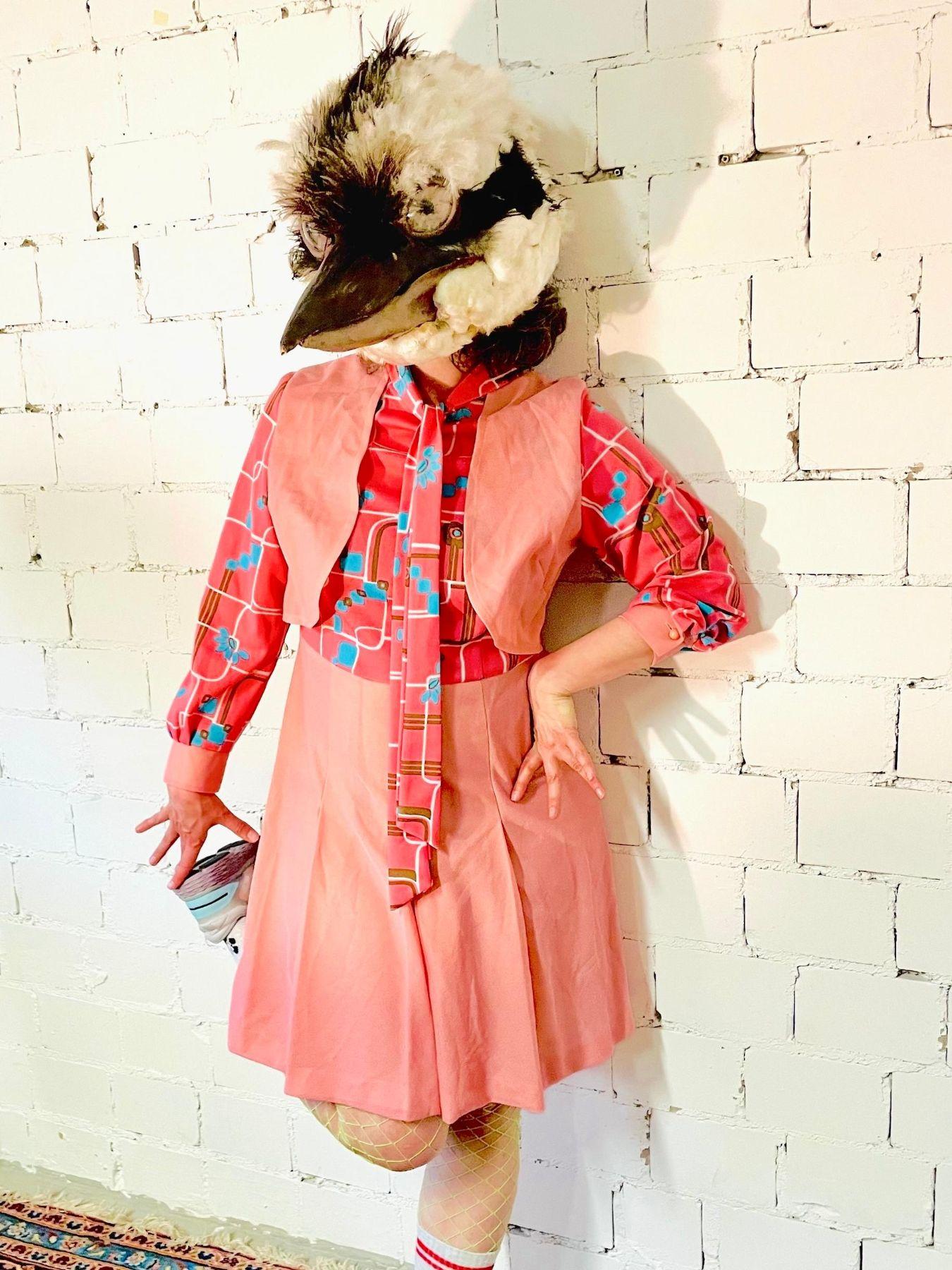 Photo of a person wearing a pink outfit. The person is wearing a bird mask and lifting one leg backward.