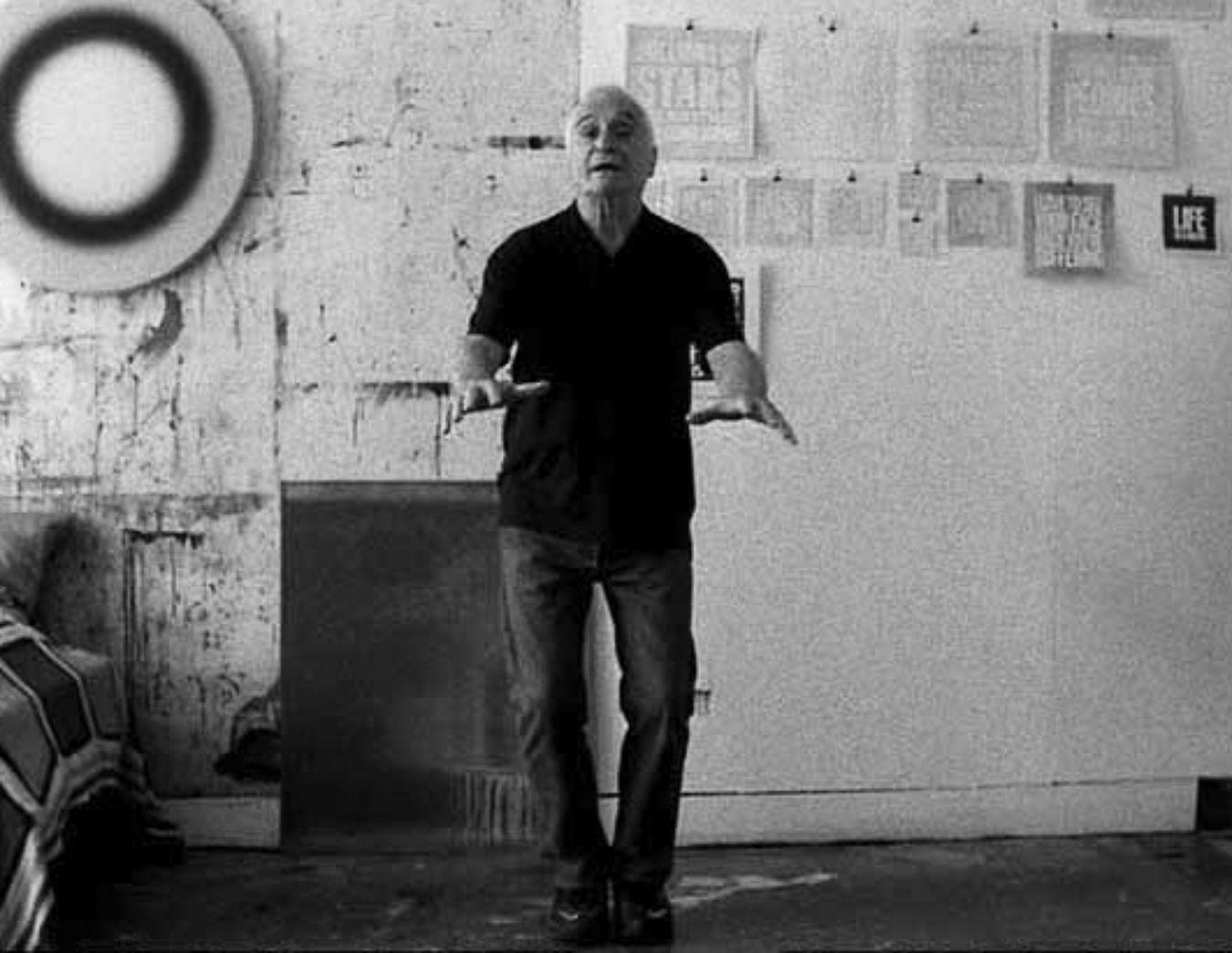 Film still from Rirkrit Tiravanija's "untitled 2008 (john giorno reads)", in which the artist John Giorno gestures towards the camera.