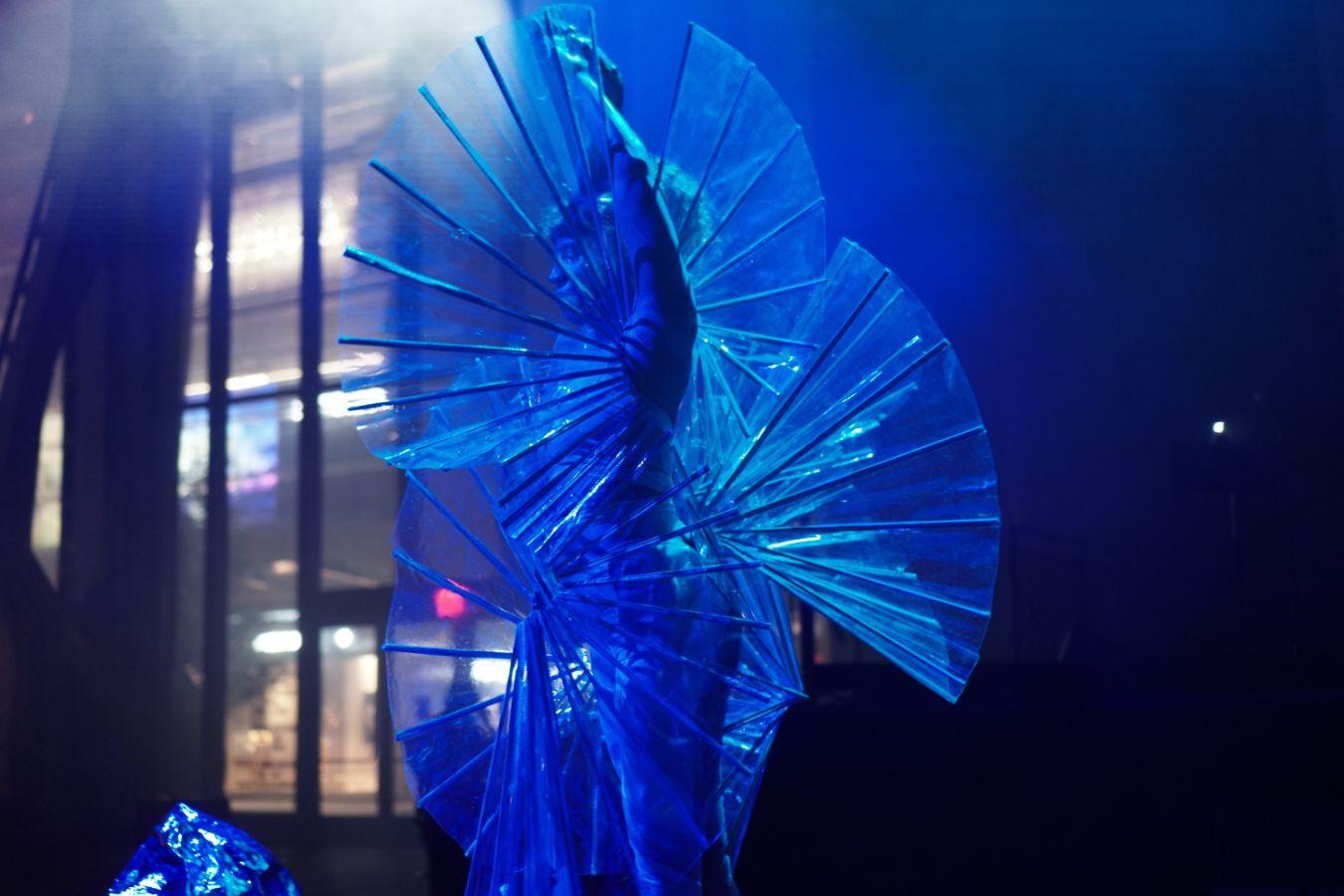 The performer Miriam Parker stands on stage wearing a fan-like costume. She holds both arms crossed over her head and is spotlit by blue lights.