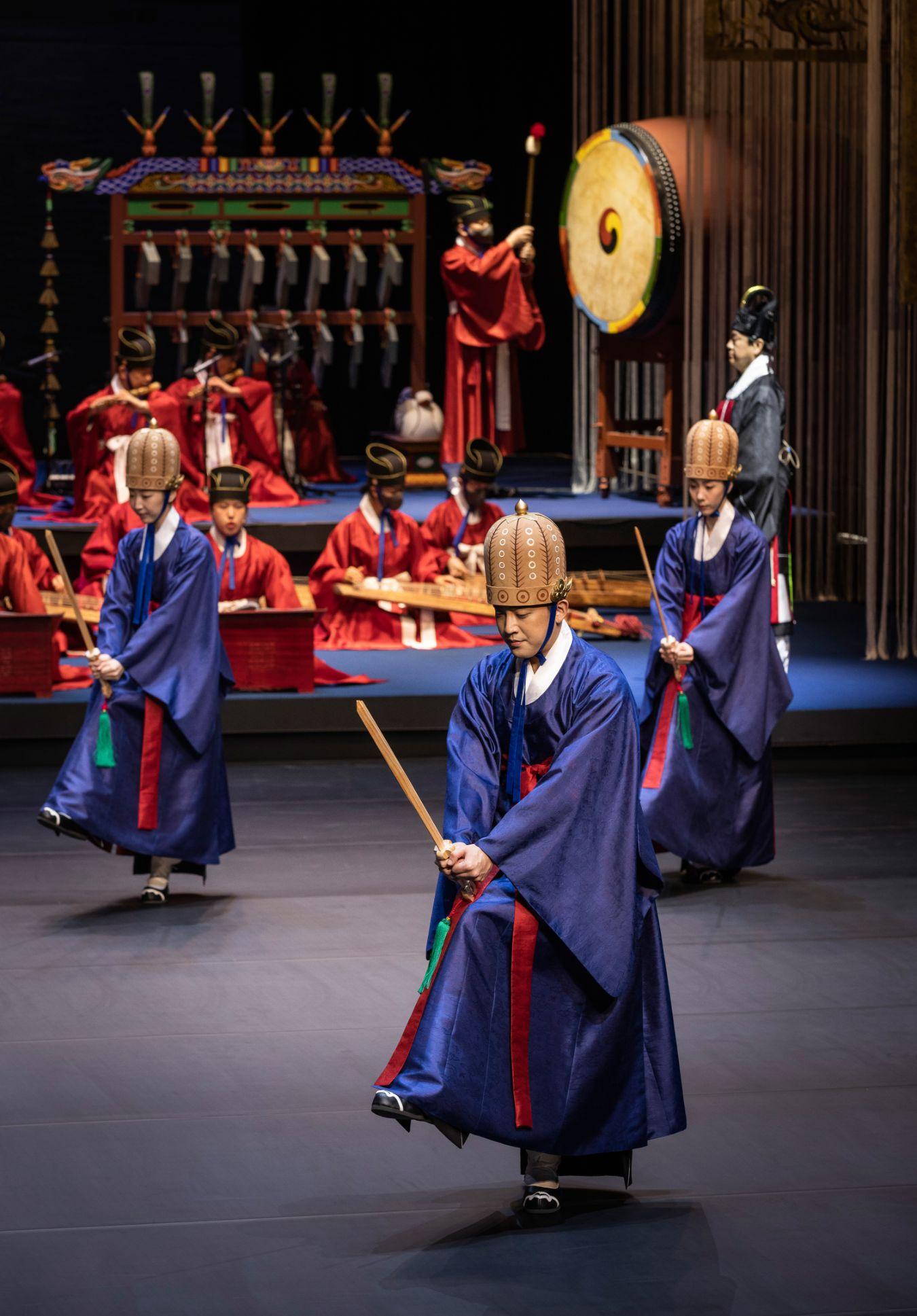 Men in ritual blue robes dance with wooden swords in their hands, behind them is an orchestra in ritual red robes.