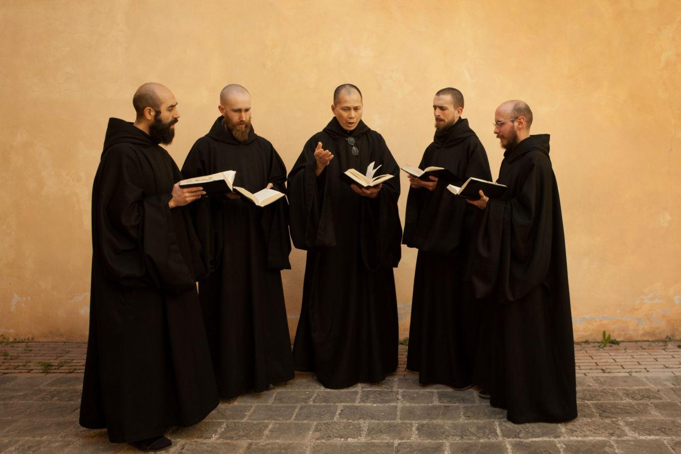 Five monks chant in a semicircle with books in their hands.