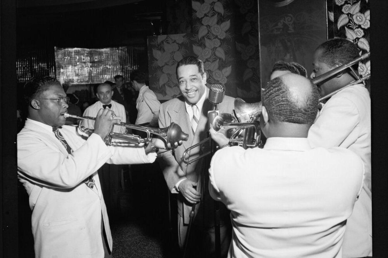 Four people with instruments play towards a man with a microphone facing the viewer.