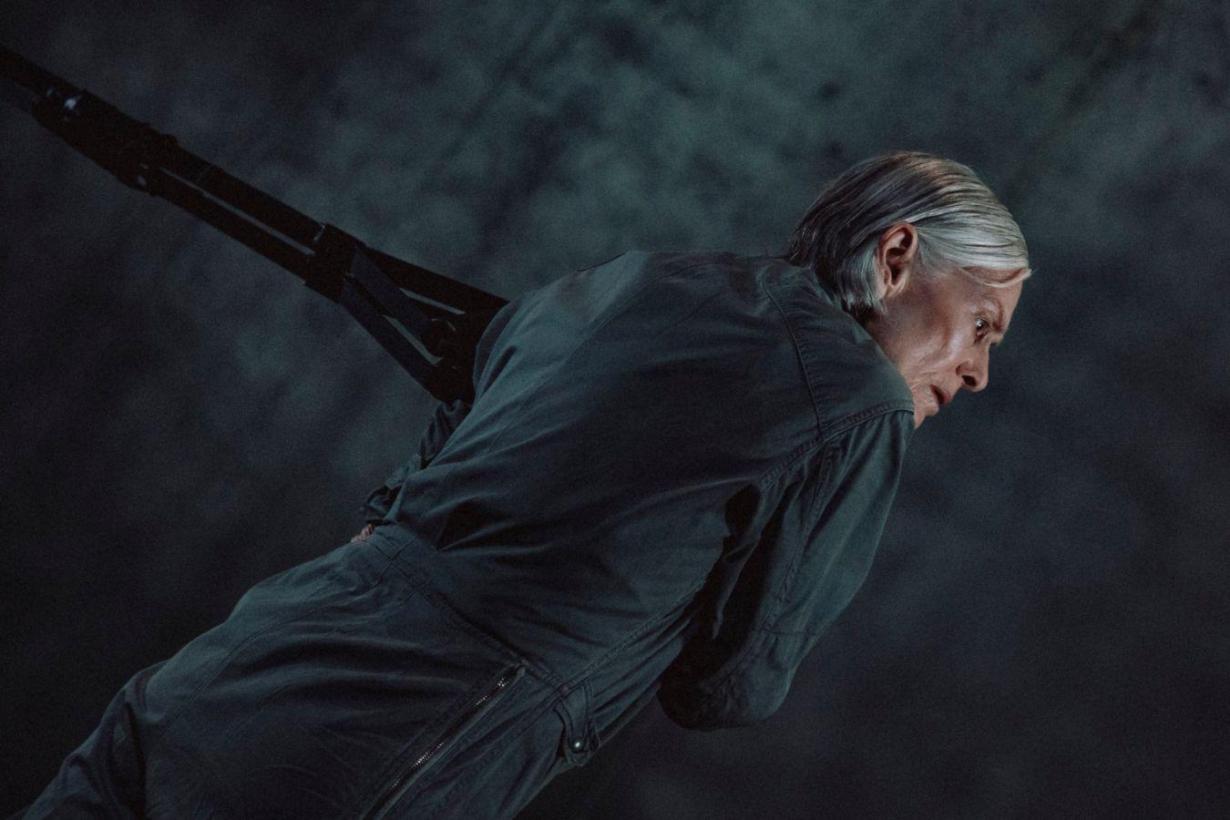 Lucinda Childs zieht an einem Seil. Die Choreografin trägt einen dunkelgrauen Overall und hat graue Haare.