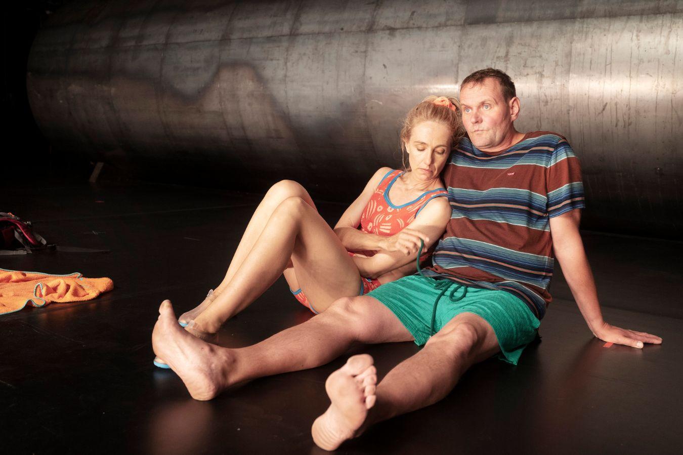 A woman and a man in summer clothes are sitting on the floor. Behind them is a huge metal pipe.