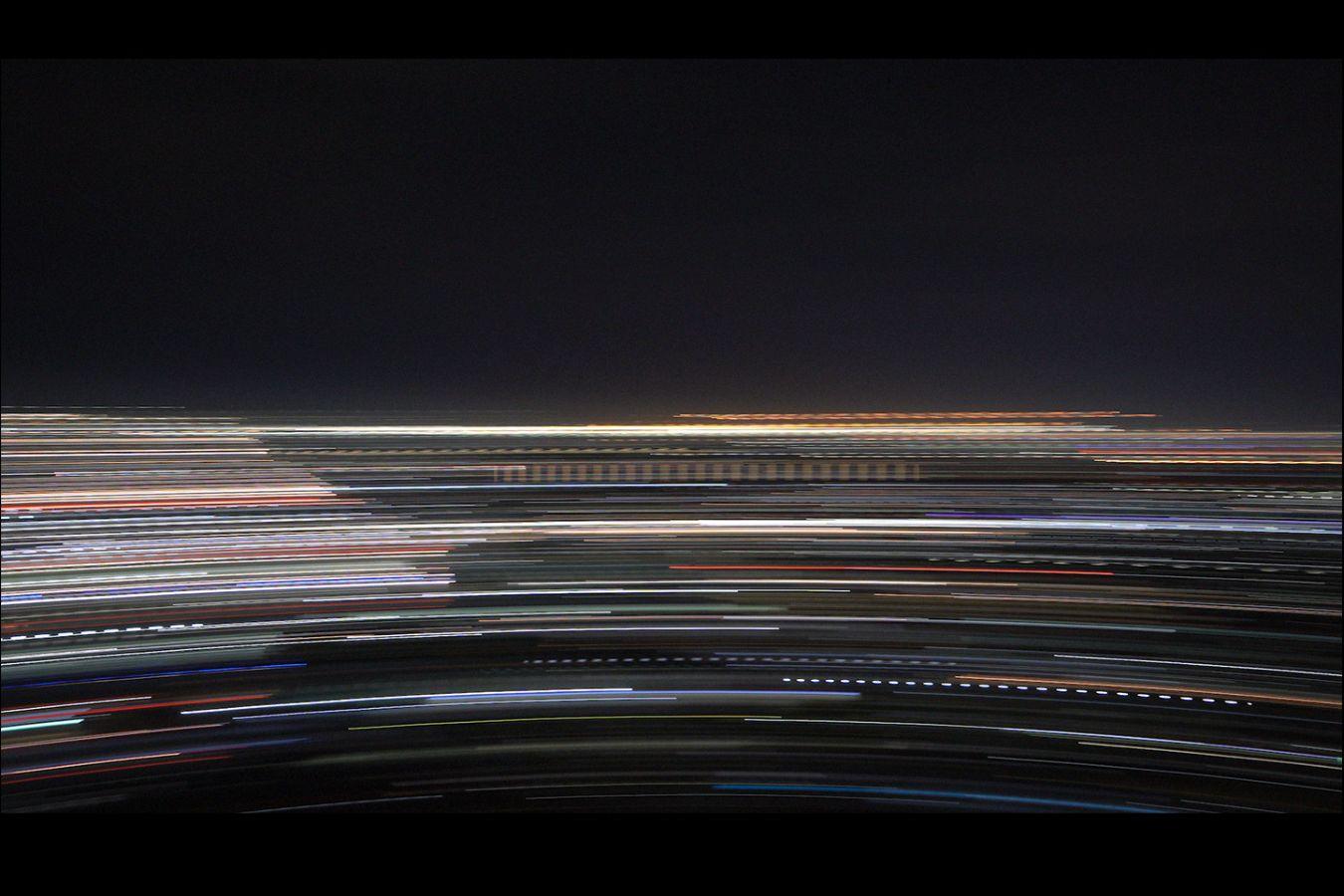 The abstract video still shows a long-exposure photography. A horizontal, fast movement creates coloured lines against a dark background in the shape of a rainbow. It invites associations, such as of a city at night with lights and traffic.