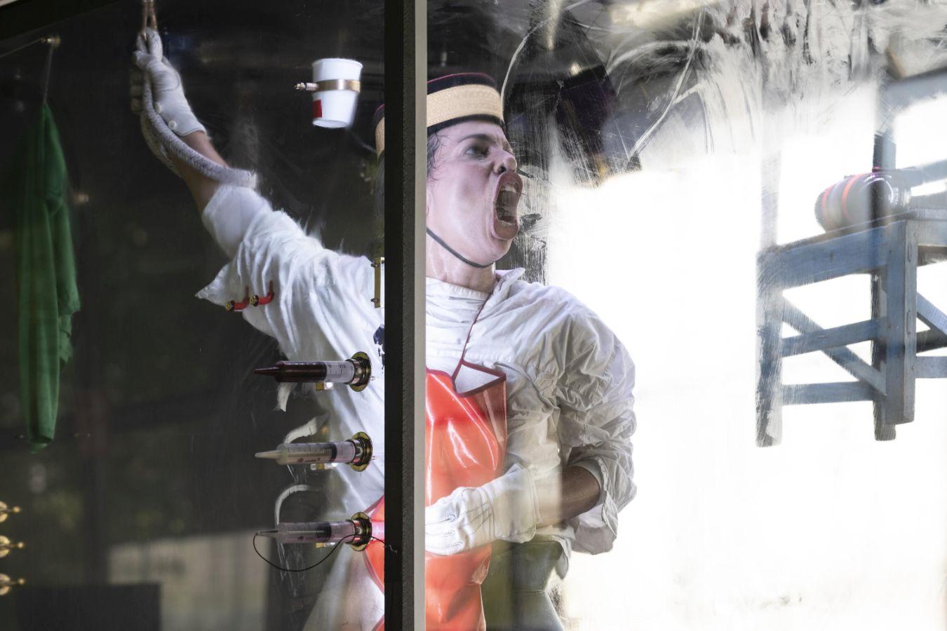 Idiota in the glass case has a red apron and white gloves on and is pressing his open mouth and his nose flat against the glass. A cup is fixed at head height. Beneath it are three syringes drawn-up with coloured liquid.