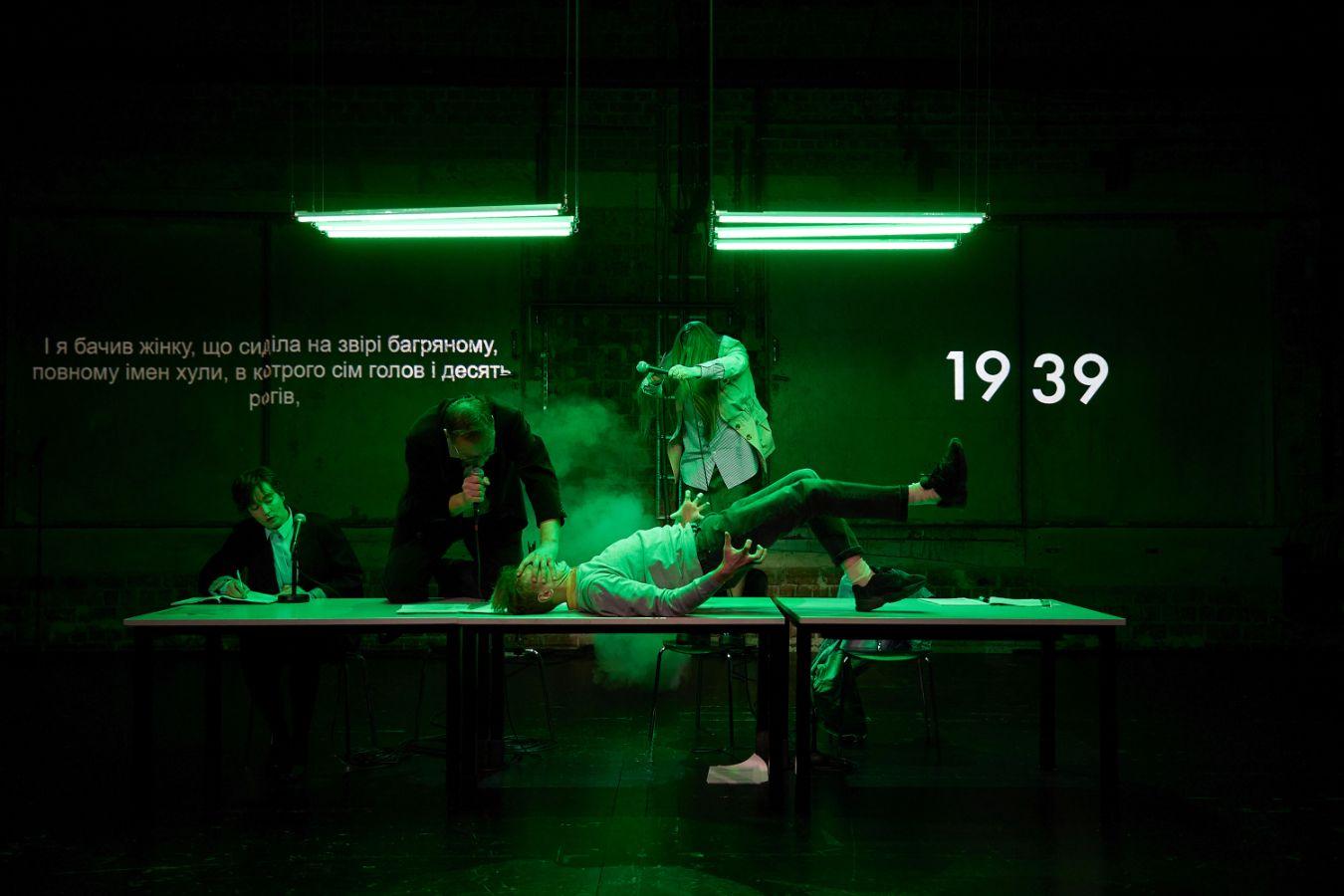 The stage glows in green neon light. In the background the number 1939 in the foreground a long table: one person speaks into a table microphone, one climbs a ladder and one person pushes down the face of another person on the table.