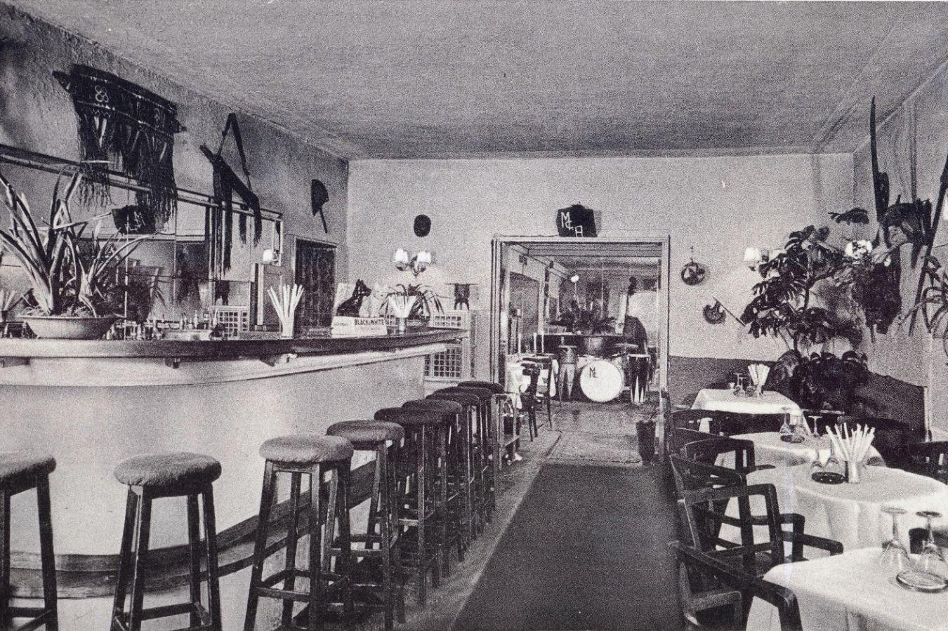 A still deserted, tidy bar with many plants and wall decorations. In the front on the left: a counter with bar stools. On the right: tables with place settings. An opening to the next room reveals instruments waiting for the band.