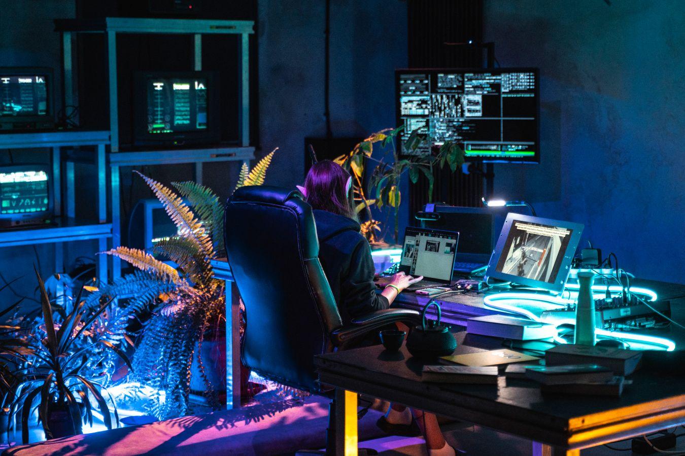 A woman with elf ears sits with her back to the camera at a desk with two screens. Around her are plants and lots of technology.