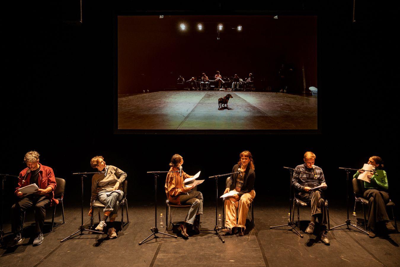 Six people sit in front of a projection of themselves and a dog