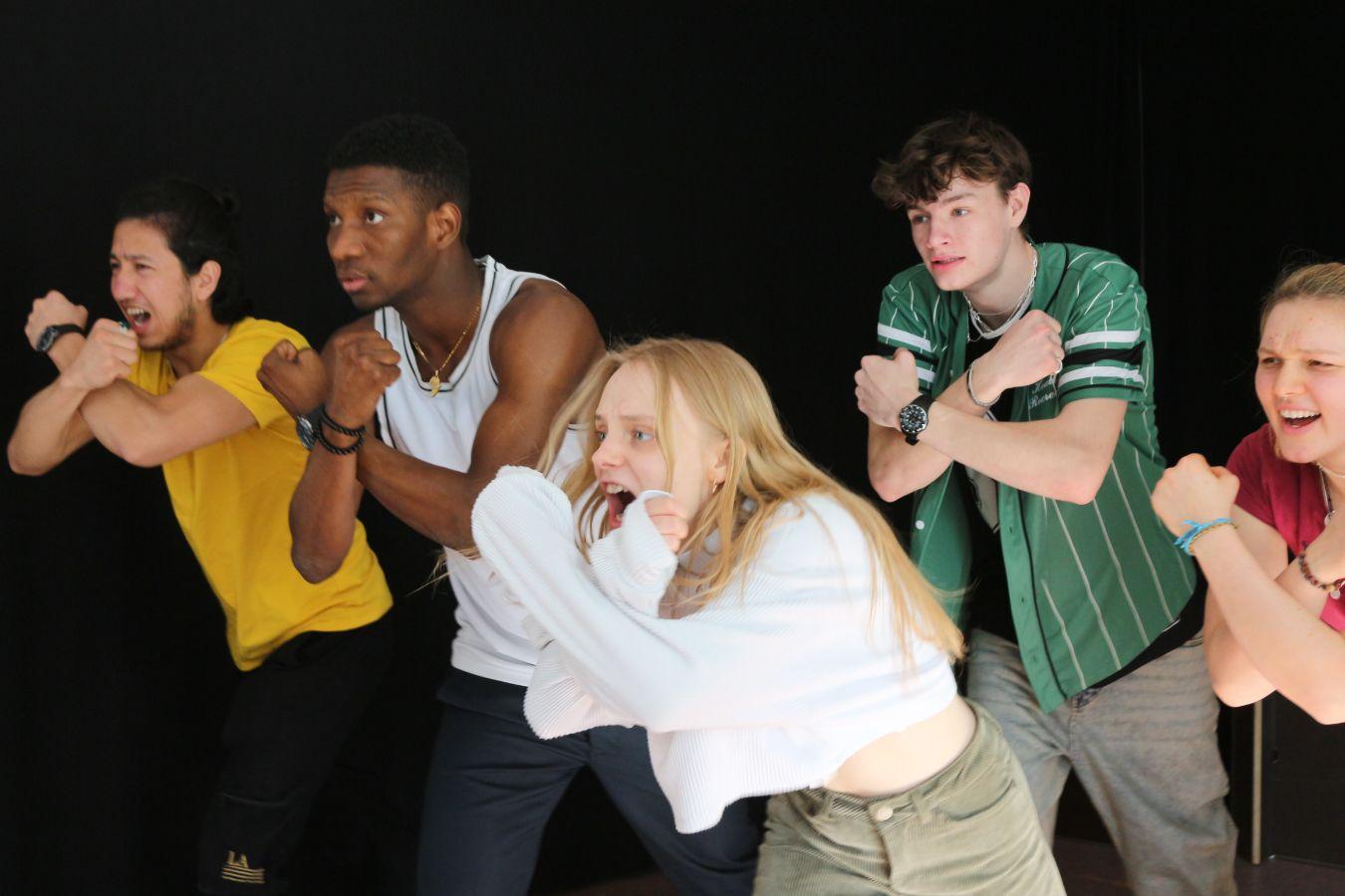 Five people are shouting loudly and have formed their arms into an X in front of their bodies. They look powerful and are wearing everyday clothes.