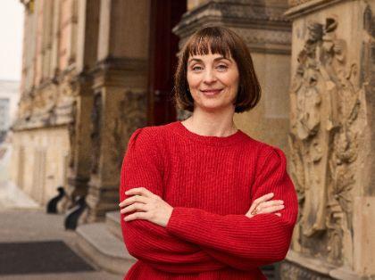 Jenny Schlenzka, dressed in a red costume, stands smiling with her arms folded in front of the entrance to the Gropius Bau.