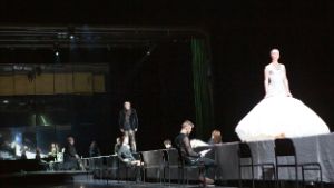 A woman in a white sweeping dress strides across a long table with people seated to the right and left.