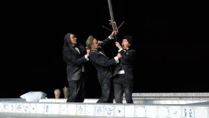 Three people in black suits stand in a tiled pool and fight over a sword.