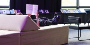 A person is lying on a modular sofa element and is reading a book. There are more books on a shelf behind the person.