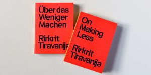 Zwei leuchtend rote Bücher liegen auf hellgrauem Hintergrund, auf ihnen steht in dunkelblauer Schrift gedruck "Über das Weniger machen. Rirkrit Tiravanija" und "On Making Less. Rirkrit Tiravanija".